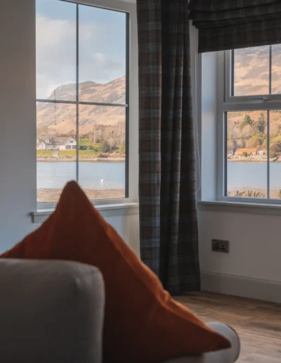 An Dòbhran atrium with grey armchair, orange velvet cushion, Harris tweed curtain and roman blinds and wide view onto the sea and the hills on the other side