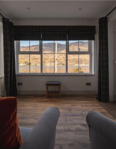 An Dòbhran atrium with grey armchair, orange velvet cushion, Harris tweed curtain and roman blinds and wide view onto the sea and the hills on the other side