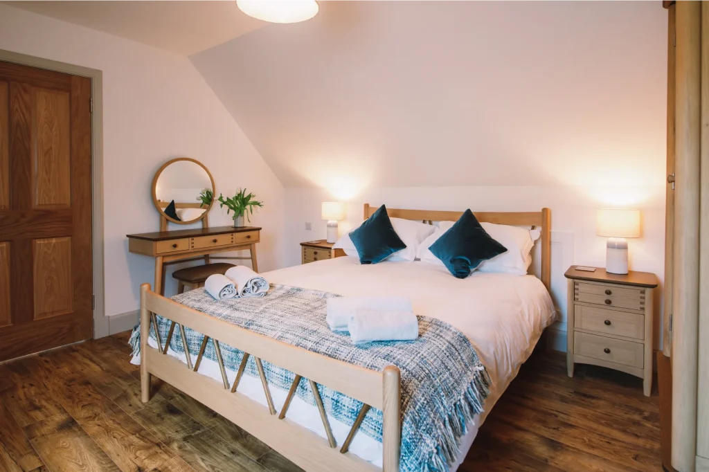 An Iolaire kingsize upstairs bedroom with wooden frame bed, matching bedside table and dressing table with mirror. Teal velvet cushion and White Company bed linen.
