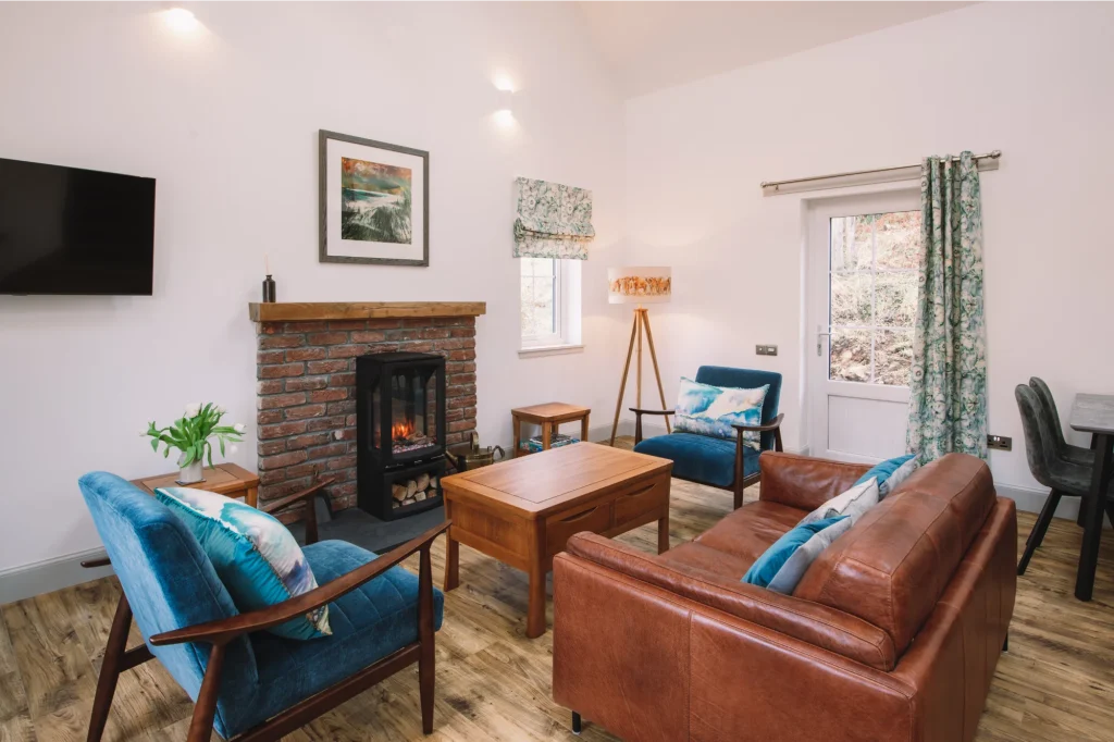 An Iolaire sitting room with side window and glazed external door. Leather couch, wood and velvet armchairs, wooden coffee and side tables, traditional brick fireplace with oak lintel and woodburner effect black stove.