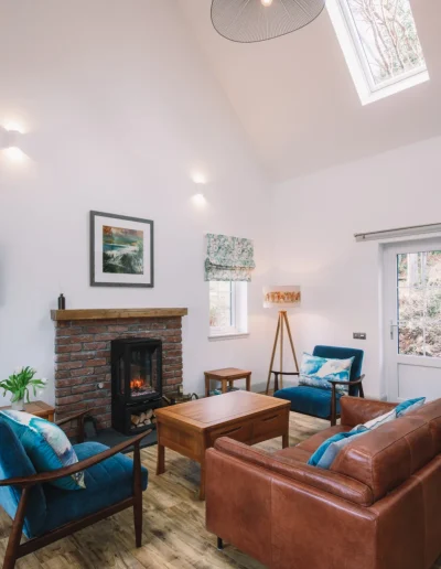 An Iolaire sitting room with vaulted ceiling and overhead skylight. Leather couch, wood and velvet armchairs, wooden coffee and side tables, traditional brick fireplace with oak lintel and woodburner effect black stove.