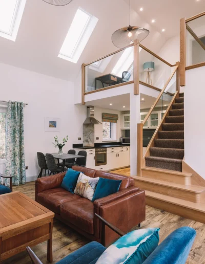 Beautiful, luxurious sitting room. An Iolaire sitting room, dining area, kitchen and stairs up to the mezzanine. Leather sofa and blue velvet armchair, highland cow cushion and lampshades, vaulted ceiling with japanese influenced black wire lightshades.