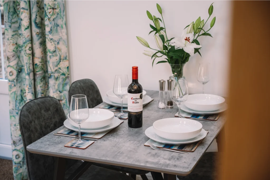 An Iolaire dining area with silver grey table, and 4 dark grey dining chairs. Table is set with crockery and glasses and someone is pouring a glass of red wine.