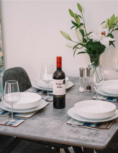 An Iolaire dining area with silver grey table, and 4 dark grey dining chairs. Table is set with crockery and glasses and someone is pouring a glass of red wine.