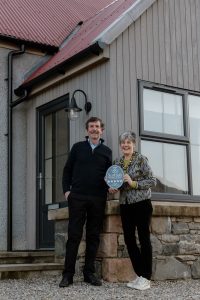 A close-up of Duncan and Shona standing in front of one of the cottages and holding their 5-star grading award from VisitScotland. Grey wood on the porch behind them, stonework by Duncan and the red corrugated iron roof.